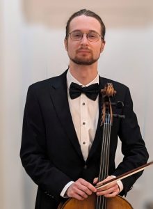 Boet is wearing a black tuxedo and bowtie, holding a cello bow with a cello.
